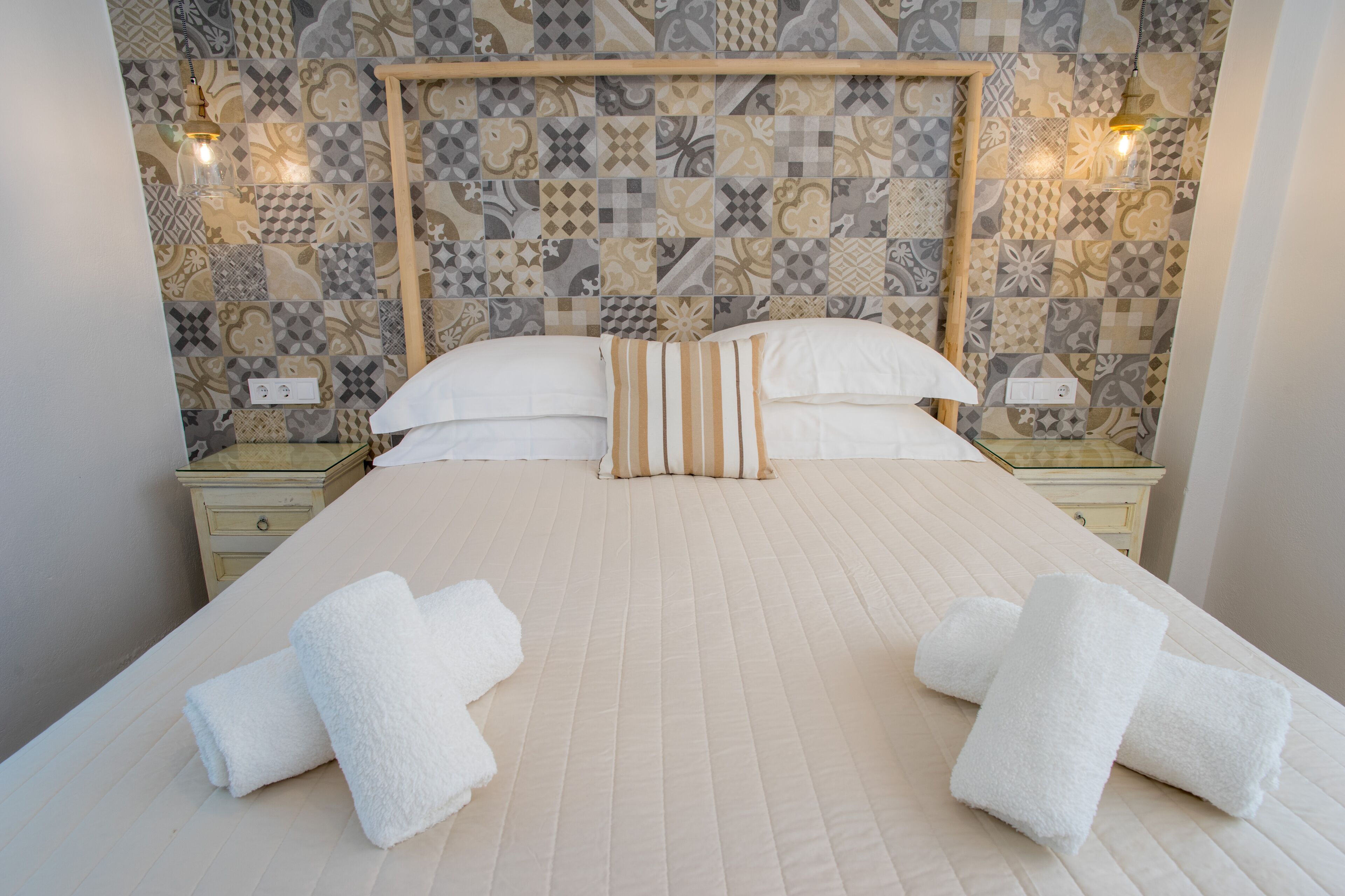 A neatly made bed with white linens and striped pillows, a wooden headboard, and a patterned tile wall.