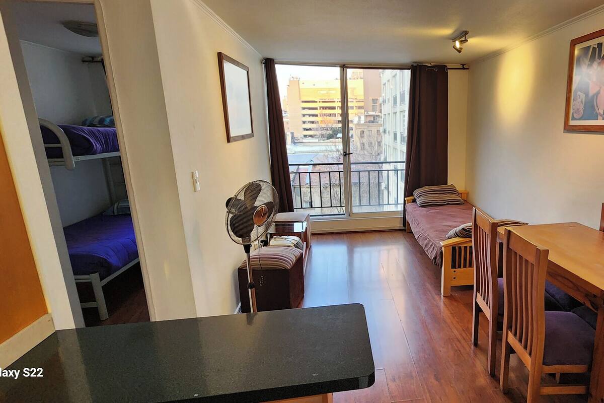 A room with bunk beds, a dining table with chairs, a fan, and a large window with a view of buildings.