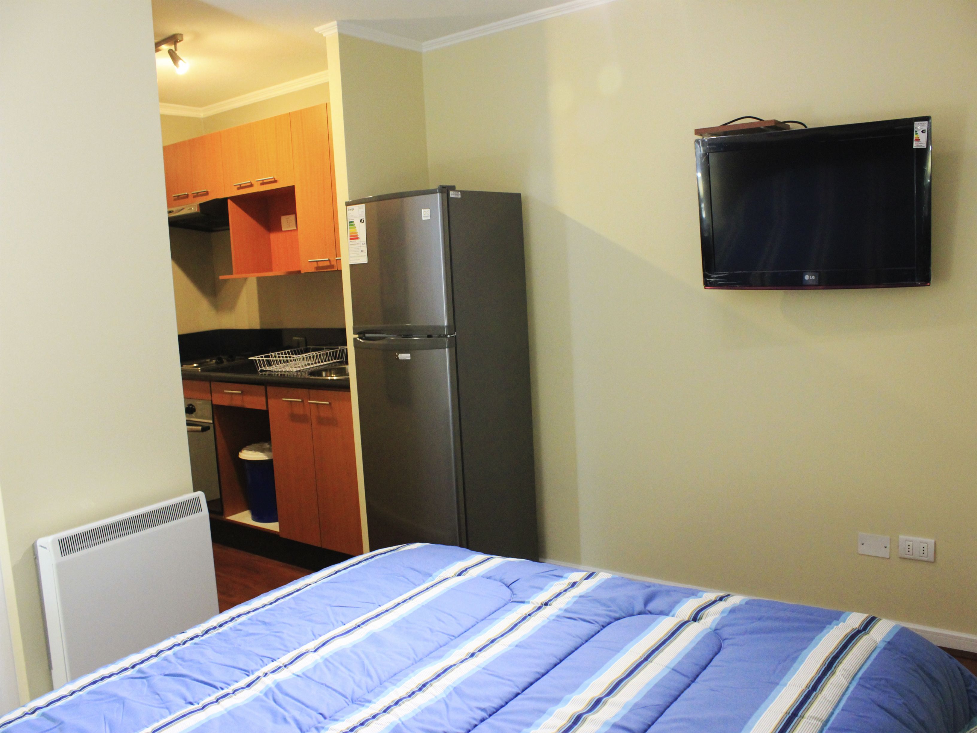 Een compacte kamer met een bed, een koelkast, een klein keukenhoekje en een televisie aan de muur.