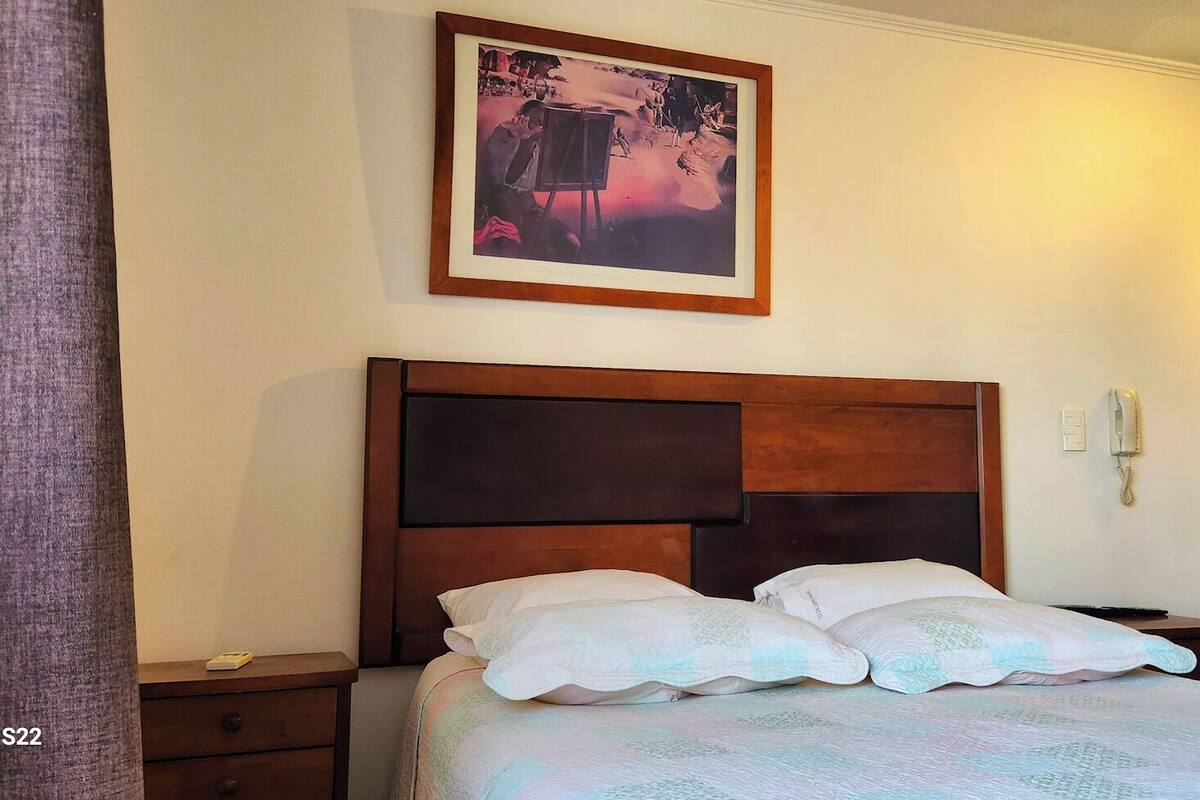 A hotel room with a large bed, a wooden headboard, a bedside table with a lamp, a framed picture on the wall, and a phone on the wall.