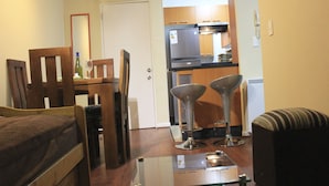 A compact living space with a bed, a glass-top table, two bar stools, a sofa, and a kitchenette in the background. - Amistar Plaza París (Santiago)