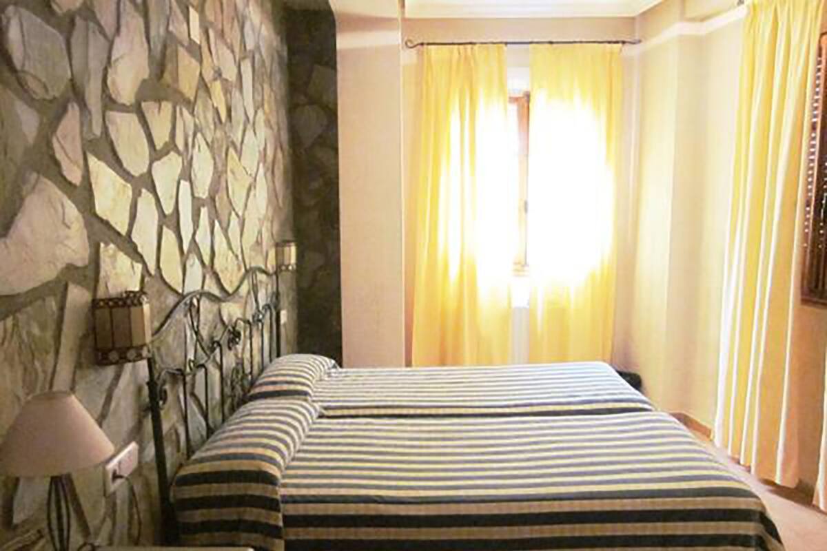 A room with a stone wall, a bed with striped bedding, a nightstand, a lamp, and yellow curtains.
