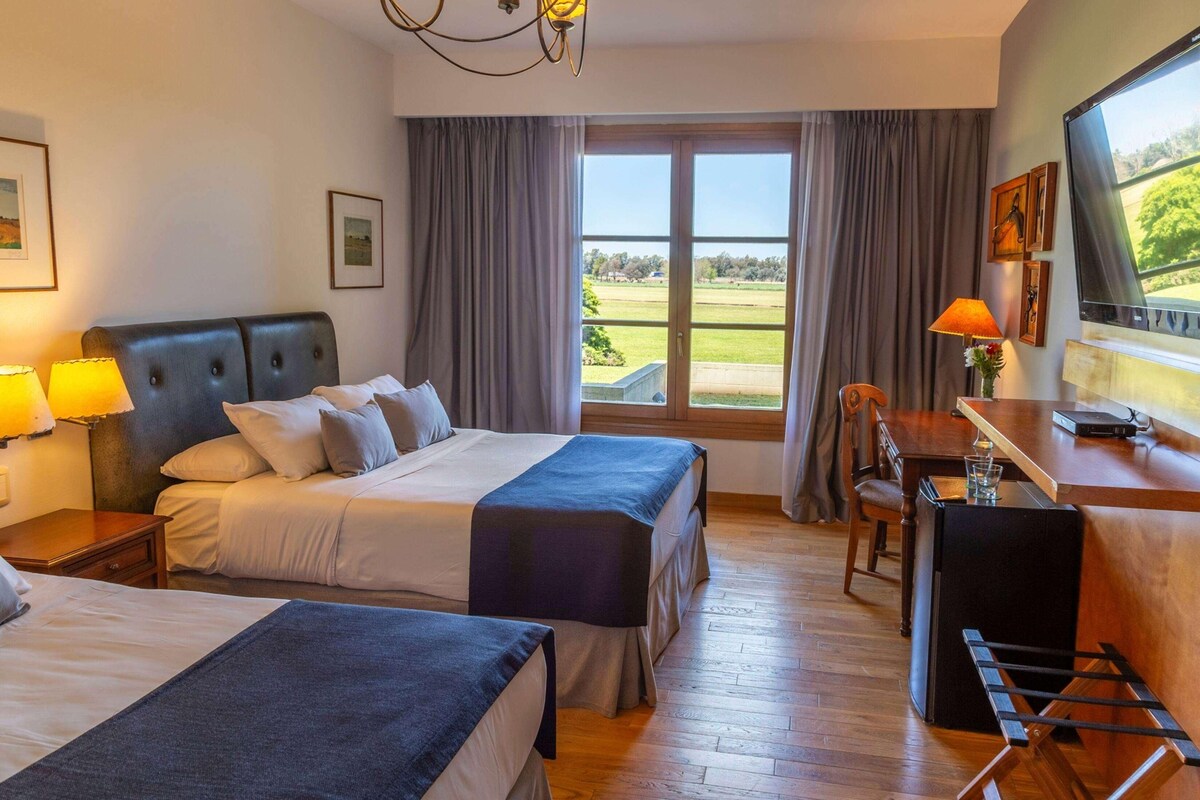 A hotel room with two beds, a desk, a chair, a TV, and a window with a view of the outdoors.
