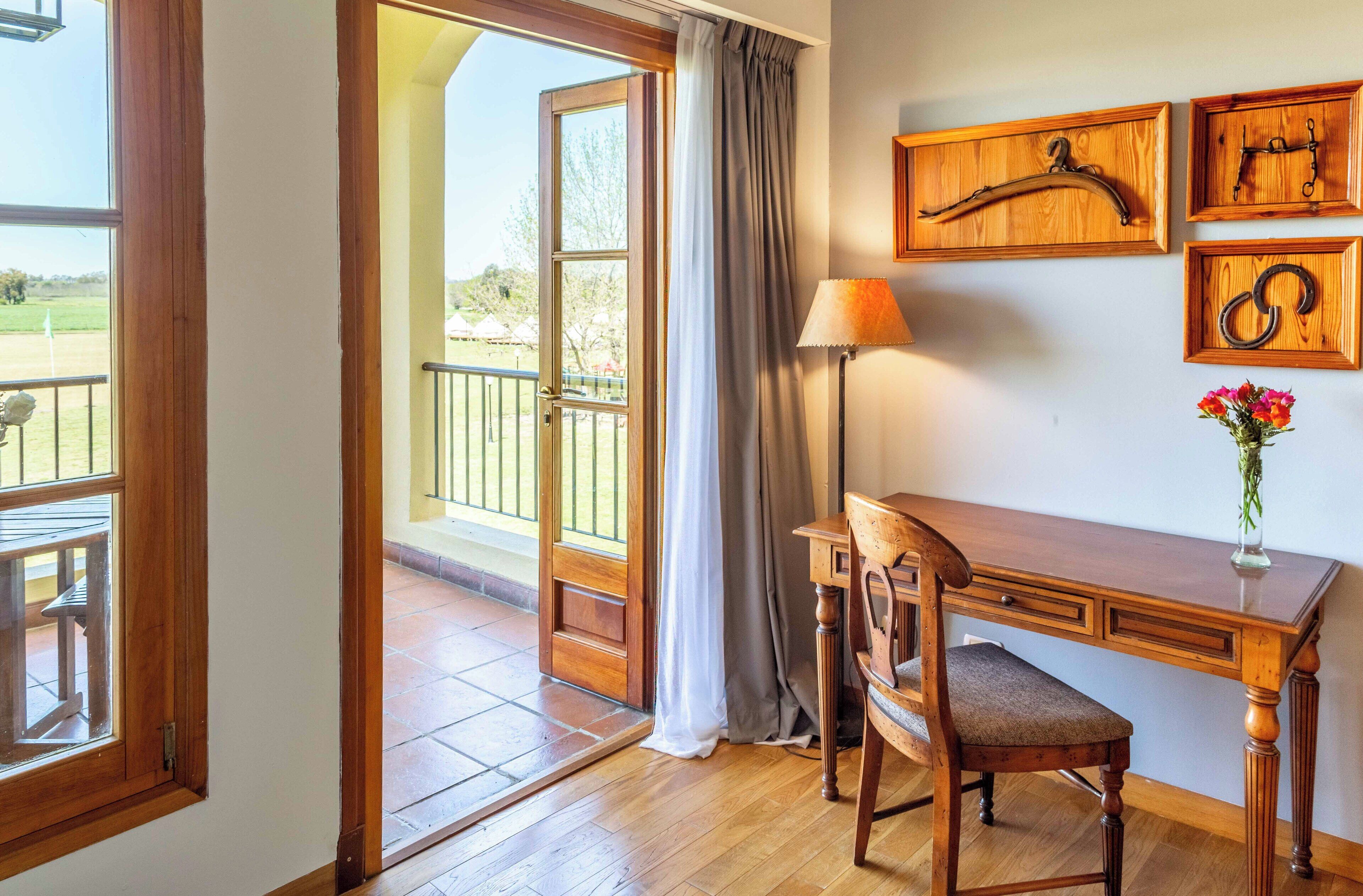 A room with a wooden desk, chair, and a view of a balcony with a table and chairs outside.