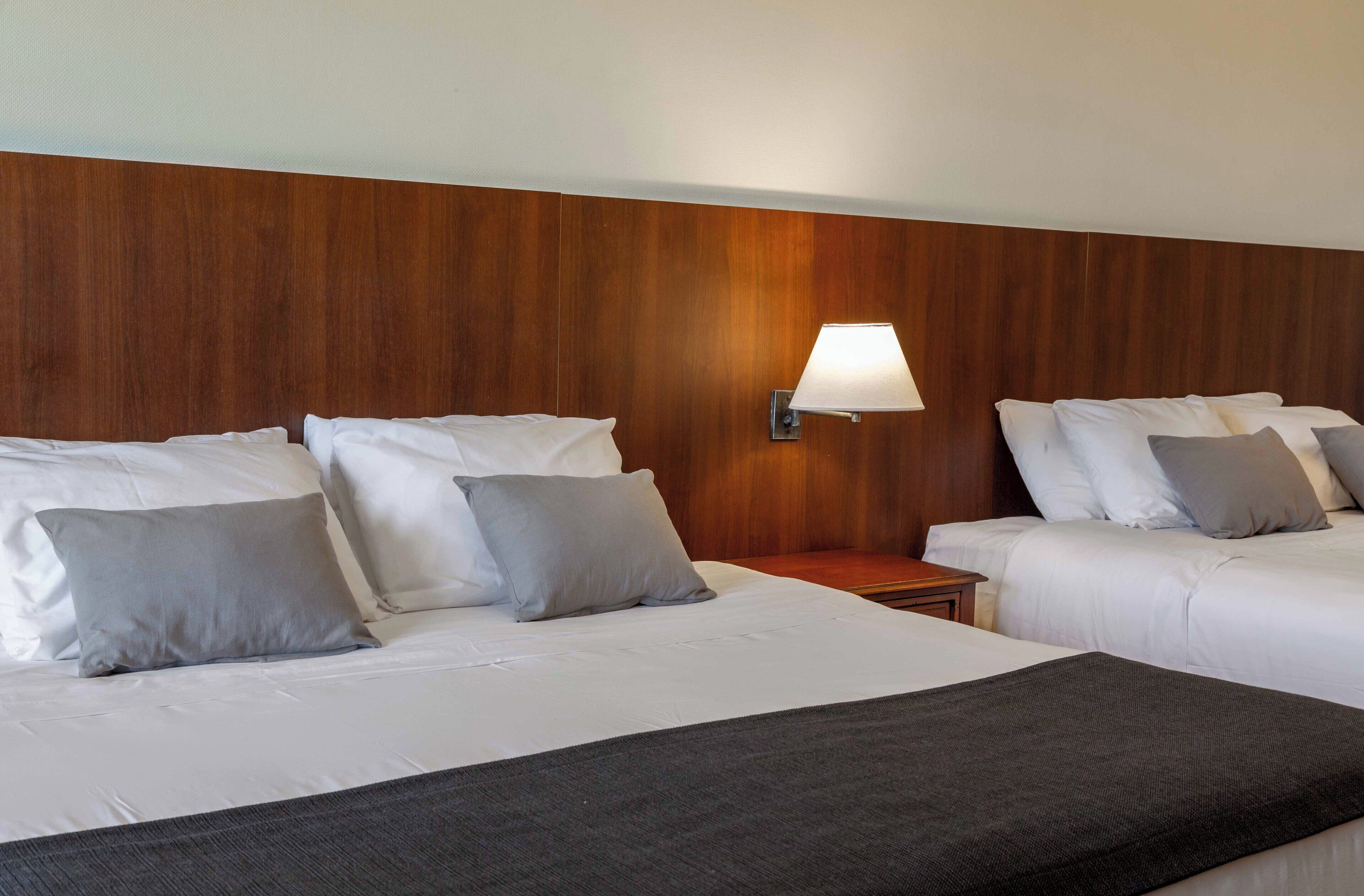 A hotel room with two beds, a wooden headboard, a nightstand, and a wall-mounted lamp.