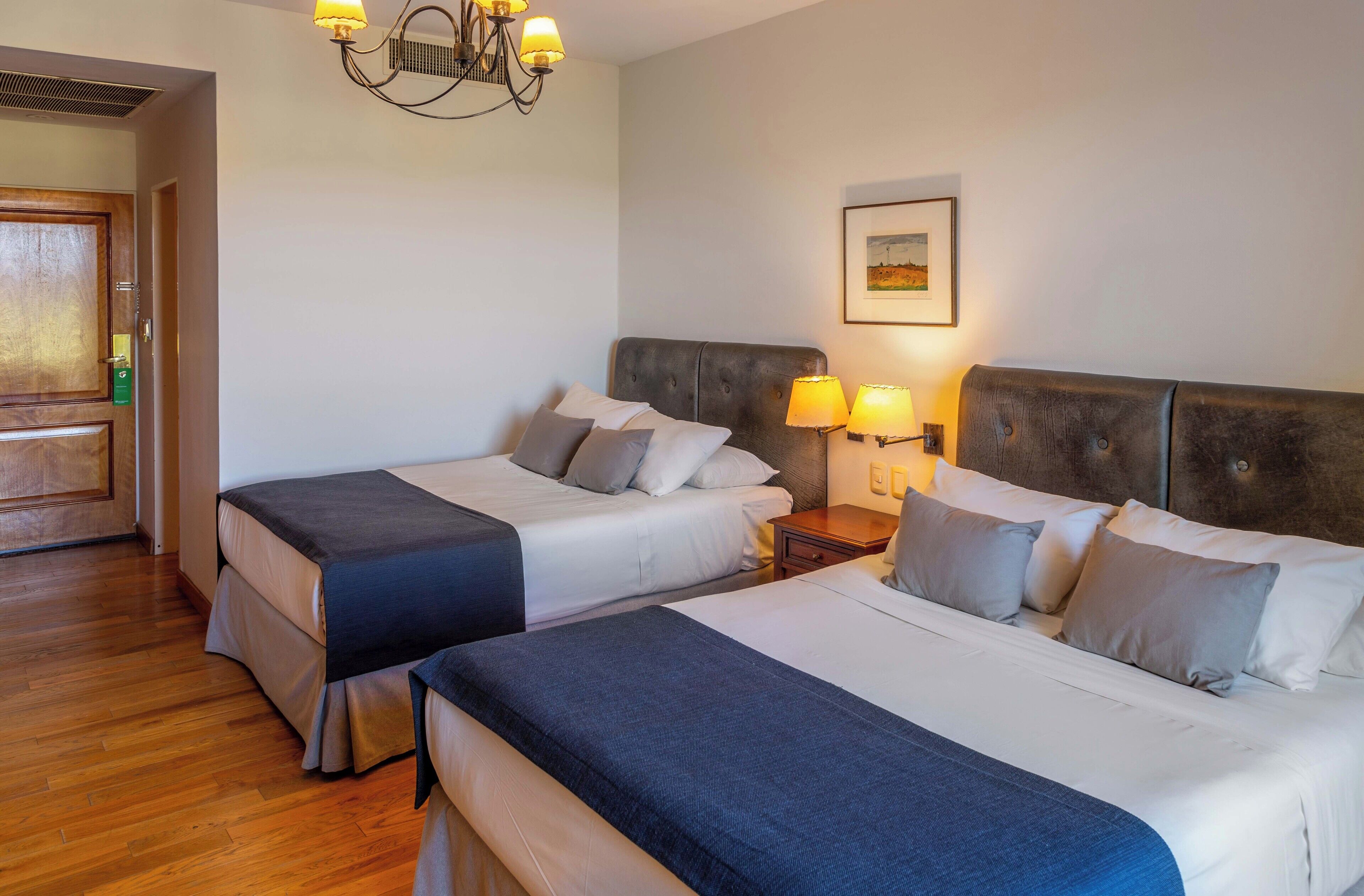 Habitación de hotel con dos camas, piso de madera, candelabro y un cuadro enmarcado en la pared.