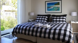 Una cama bien hecha con ropa de cama a cuadros, un cuadro de una escena de playa en la pared y vistas al verdor a través de la ventana.