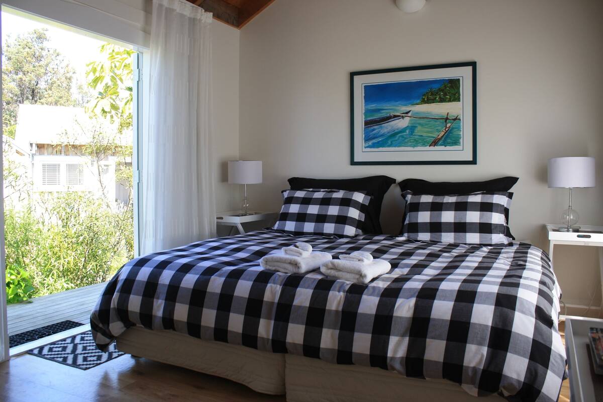 Una cama bien hecha con ropa de cama a cuadros, un cuadro de una escena de playa en la pared y vistas al verdor a través de la ventana.