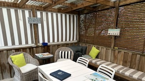 Un patio cubierto con mobiliario de mimbre, una mesa con un libro y una libreta, y un cartel con normas de la casa.