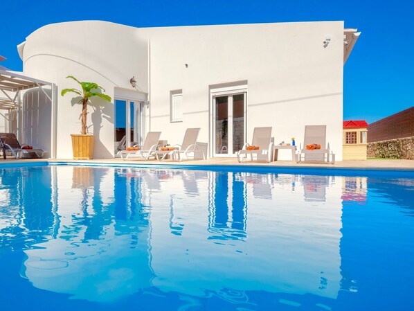 A modern white building with a pool, sun loungers, and a potted plant.