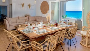 Een eetgedeelte met een houten tafel gedekt voor zes personen, omringd door rieten stoelen. Een beige bank staat tegen de muur, en de kamer heeft een groot raam met uitzicht op de zee.