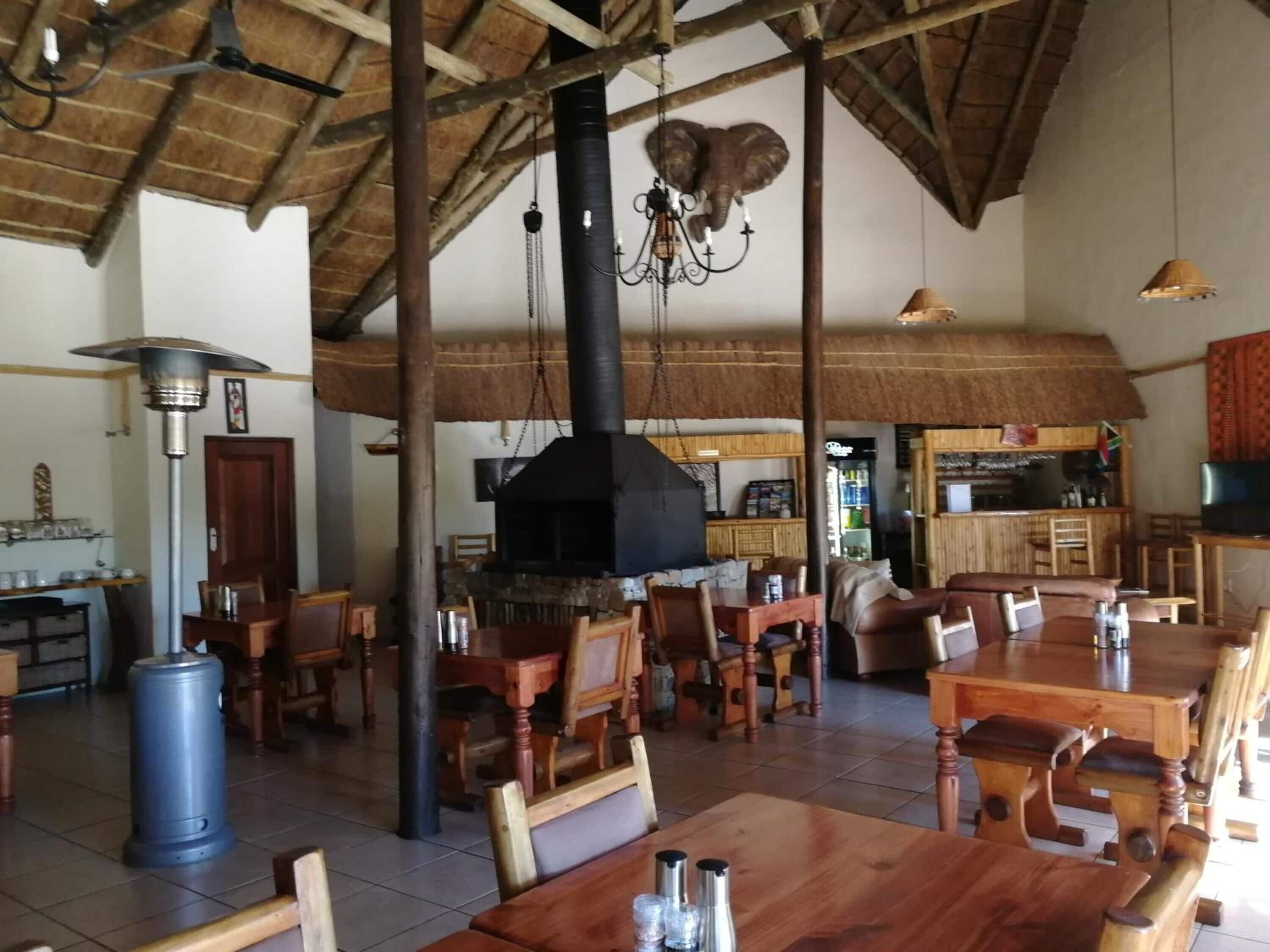 Interior rústico de un restaurante con mesas y sillas de madera, una chimenea negra grande y una escultura de elefante colgante.