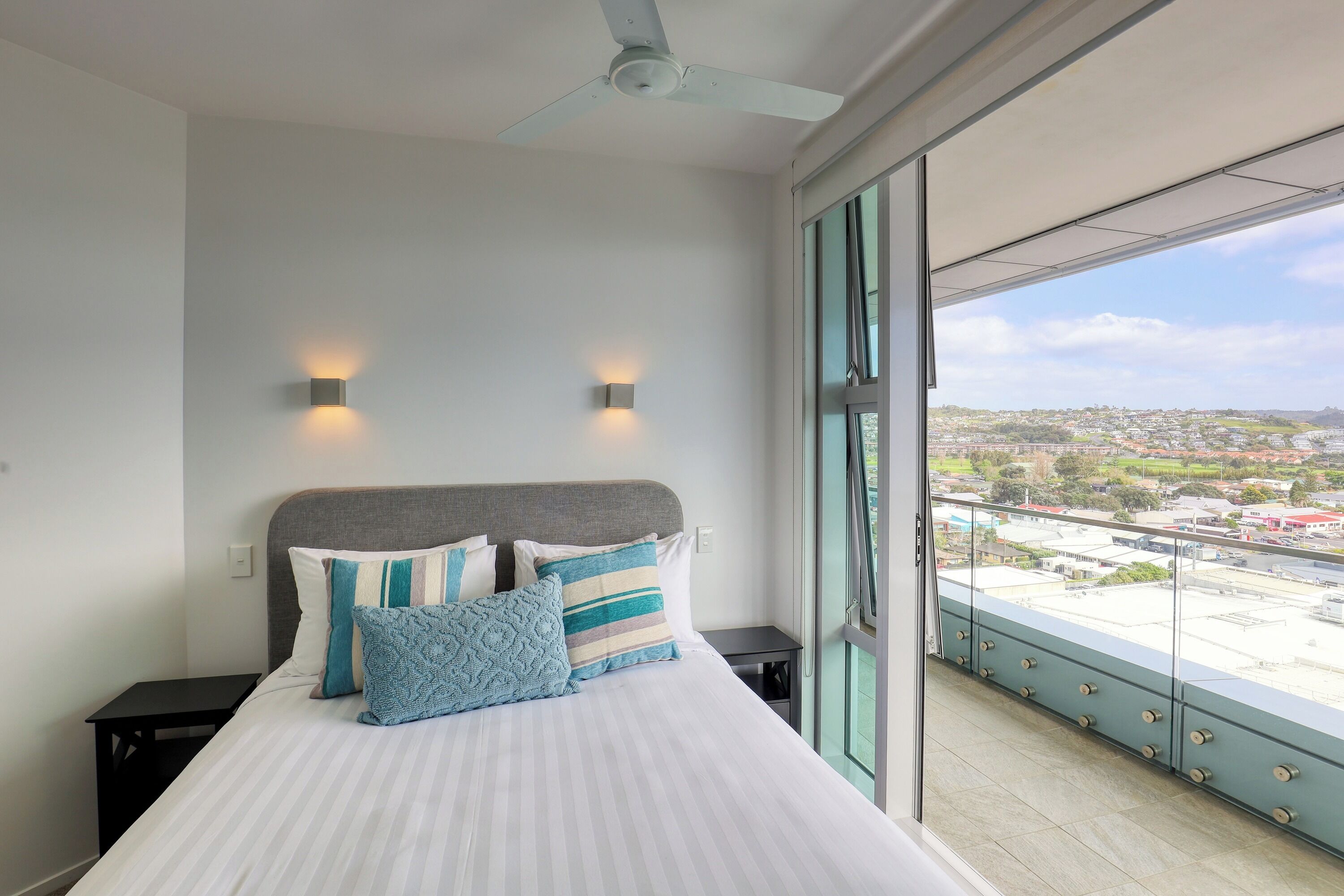 Un dormitorio con una cama grande, dos lámparas de pared, un ventilador de techo y un balcón con vista a la ciudad.