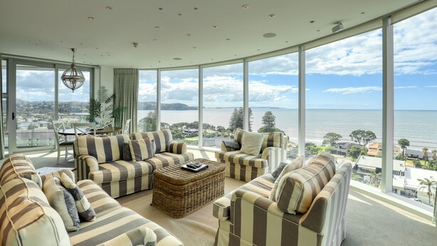 A spacious living room with striped sofas and a central coffee table, large windows offering a view of the sea and cityscape. - Marsden Suites Nautilus Orewa (Orewa)