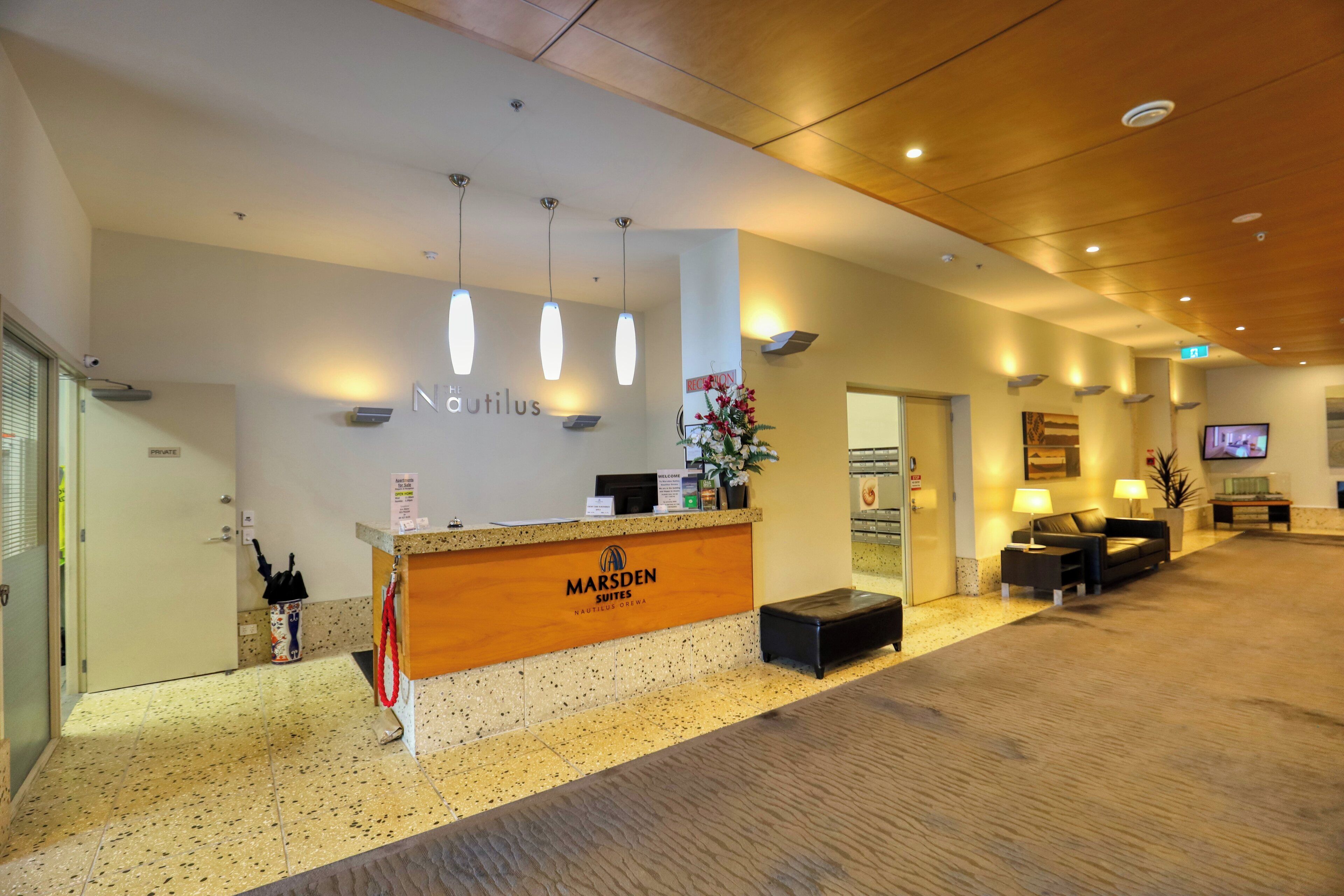 A hotel reception area with a wooden counter, a sign reading "Marsden Suites," and a reception desk. There are three pendant lights above the counter, a potted plant, and a television screen on the wall.
