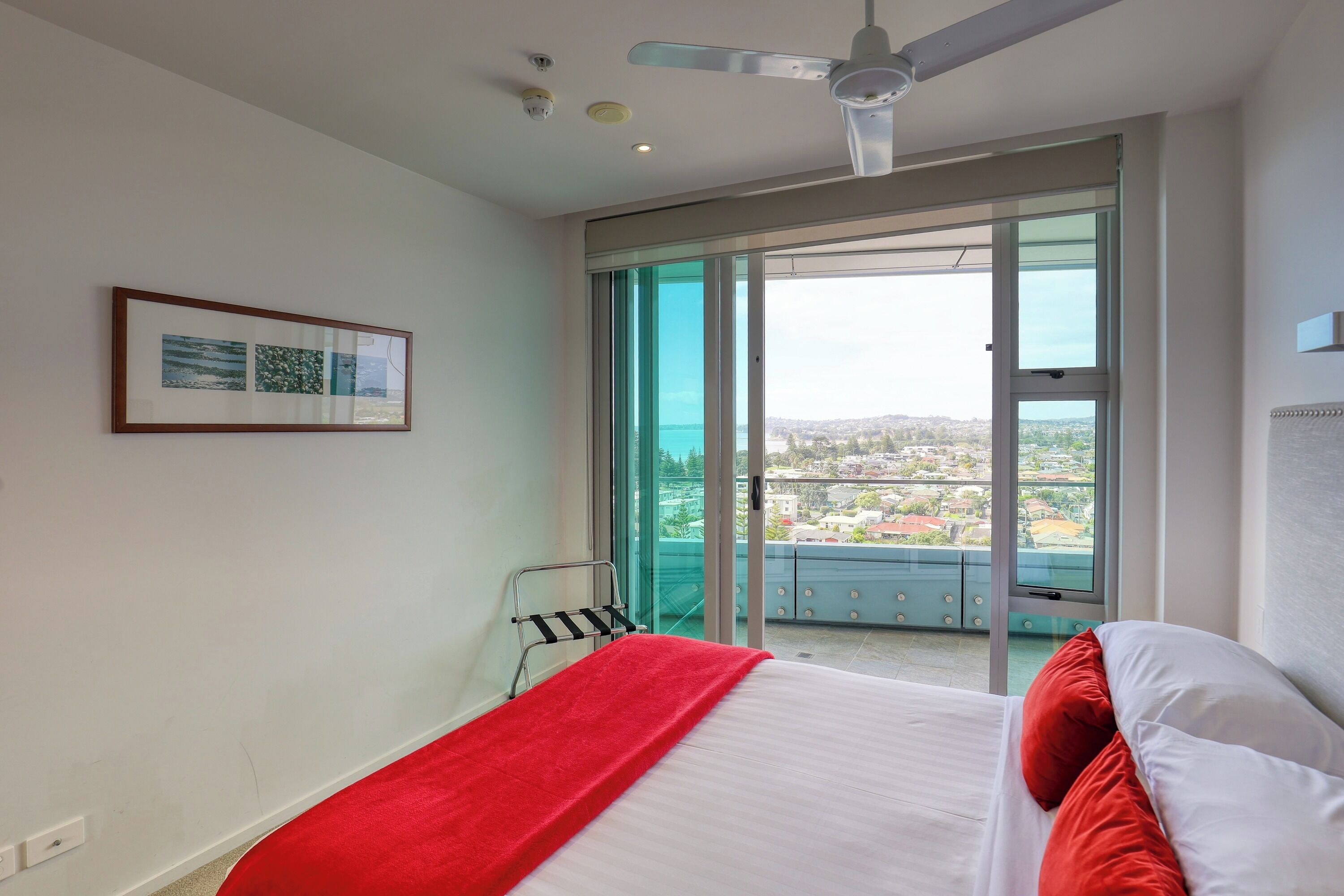 Une chambre d’hôtel avec un lit, un ventilateur de plafond, une porte-fenêtre coulissante donnant sur un balcon et un tableau encadré au mur.