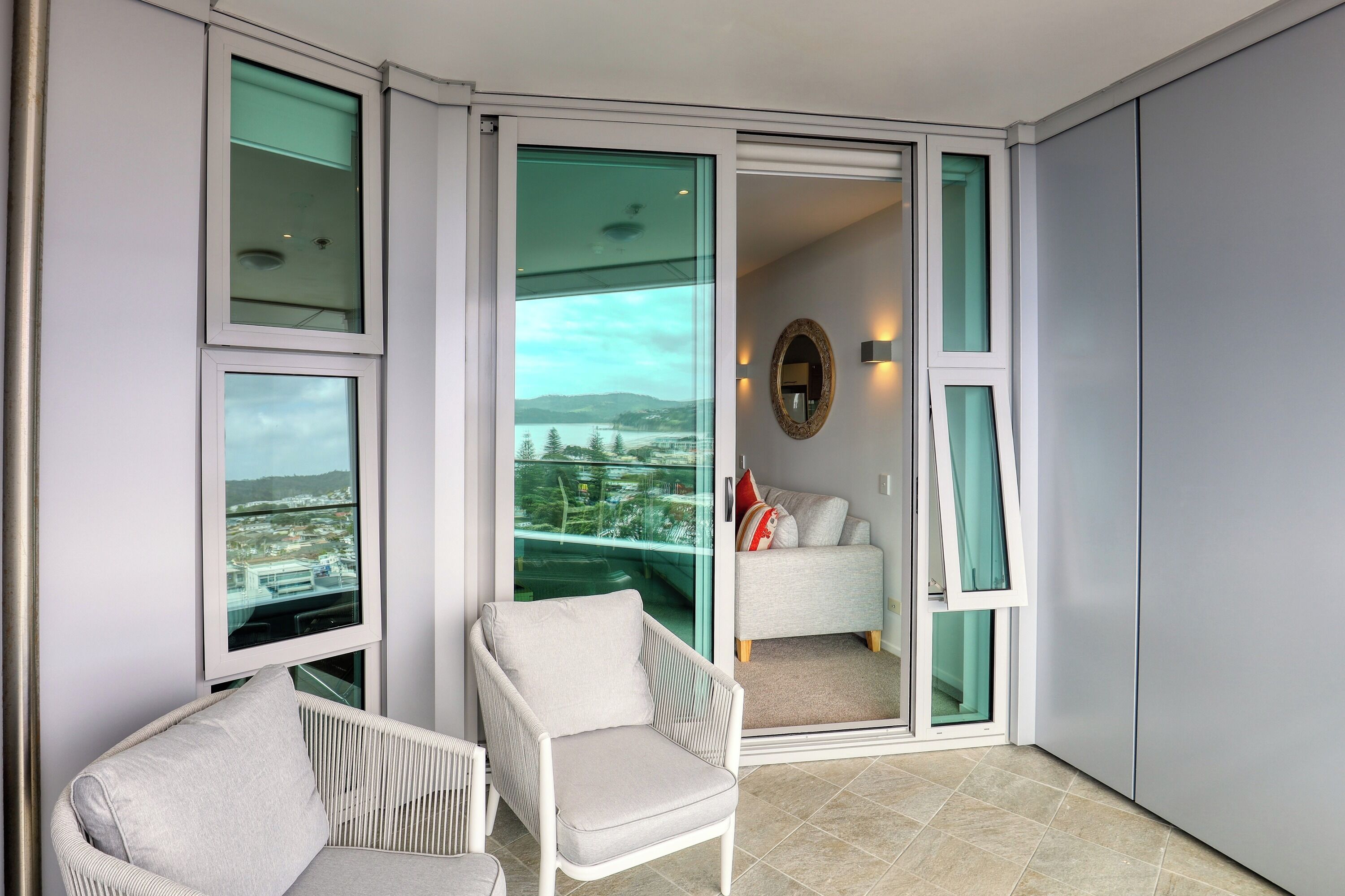 En balkon i hjørnet med udsigt over bybilledet, en hvid stol og en sofa med et spejl og væglamper.
