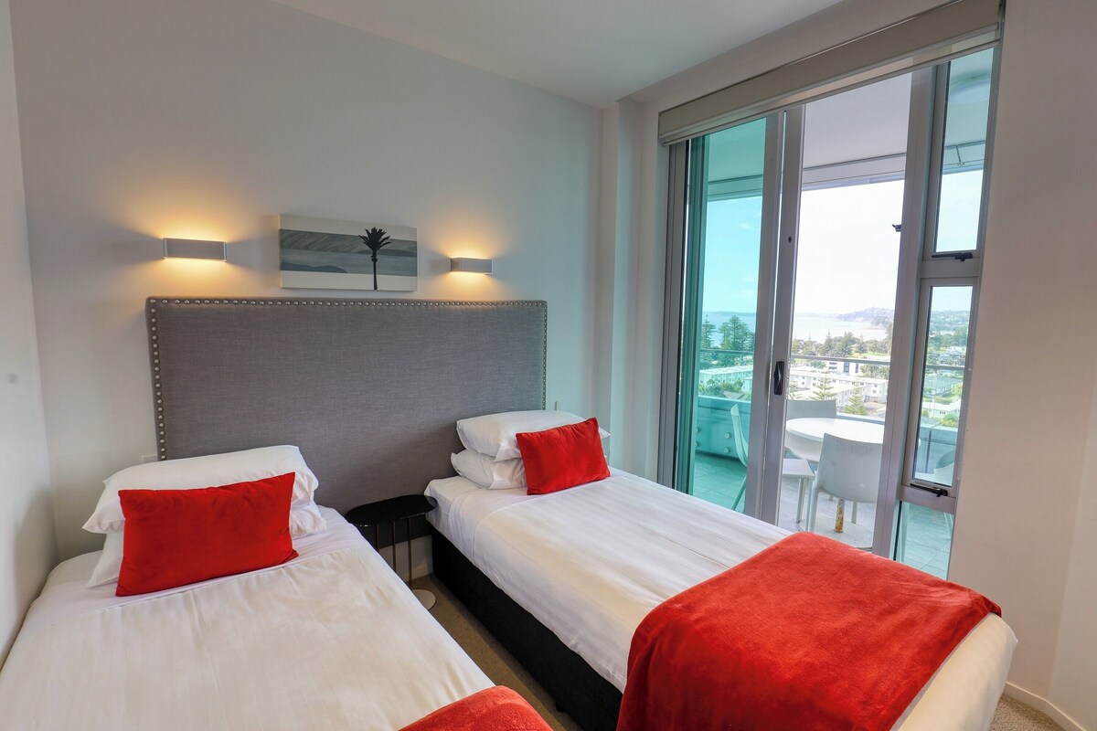 Habitación de hotel con dos camas, cada una con ropa de cama blanca y almohadas rojas, un cabecero gris y un ventanal con vistas a la ciudad.