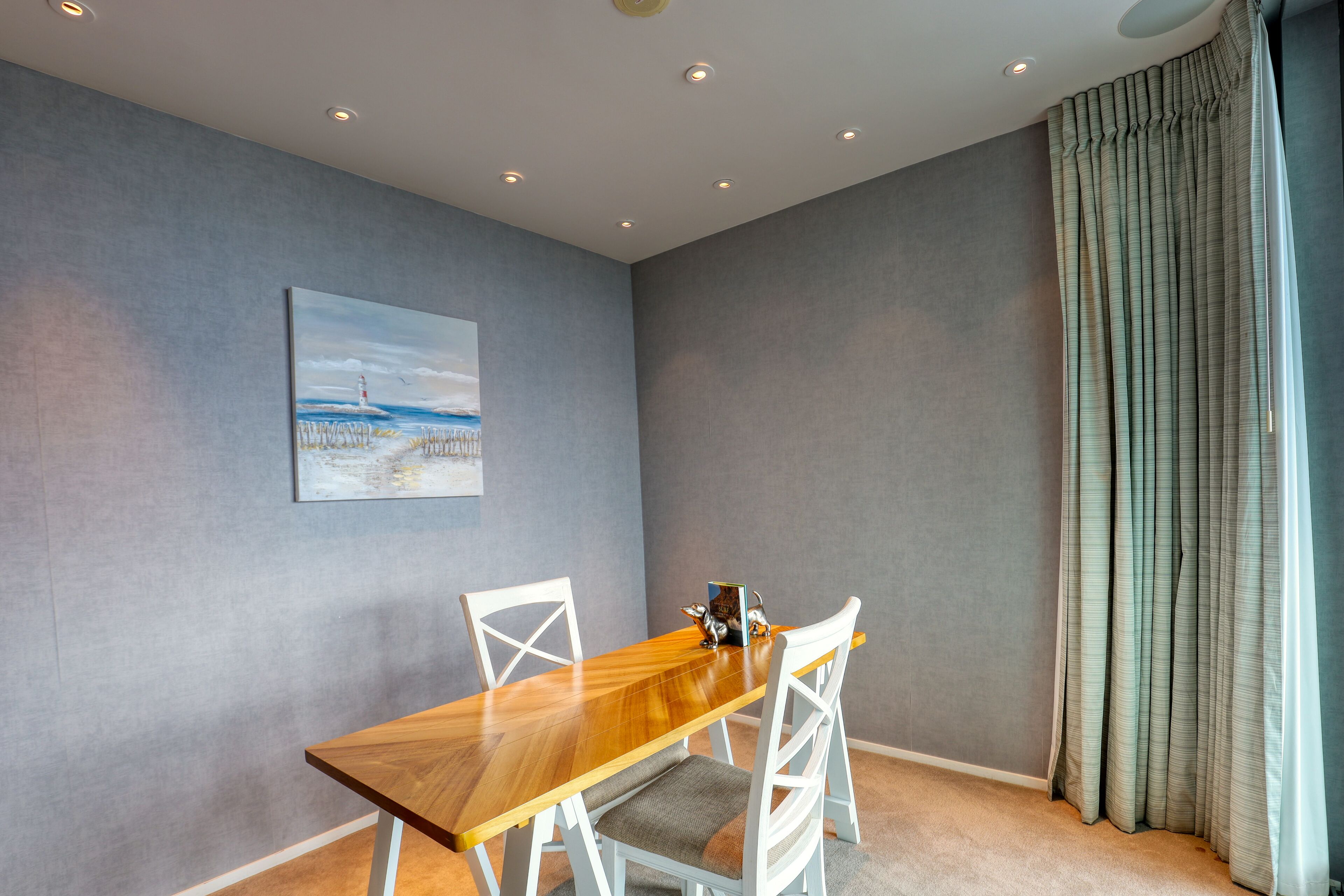 A dining area with a wooden table and white chairs, a painting on the wall, and a window with curtains.