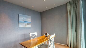 A dining area with a wooden table and white chairs, a painting on the wall, and a window with curtains.