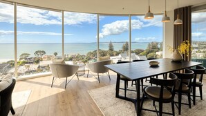 A dining area with a large table and chairs, large windows offering a view of the sea and cityscape. - Marsden Suites Nautilus Orewa (Orewa)