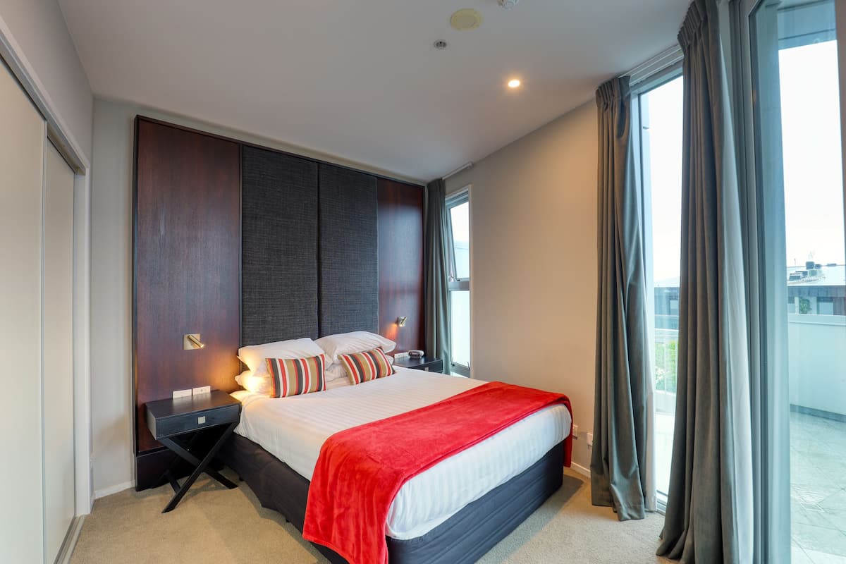 A modern hotel room with a large bed, a nightstand, a red throw blanket, and a view of the city through large windows.