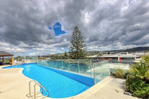 A clear blue swimming pool with a glass railing, surrounded by greenery and a cloudy sky. - Marsden Suites Nautilus Orewa (Orewa)