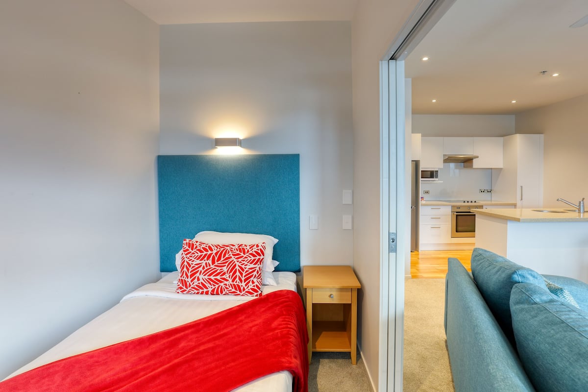 A compact bedroom with a bed, a bedside table, a red blanket, and a blue headboard. A kitchenette is visible in the background.