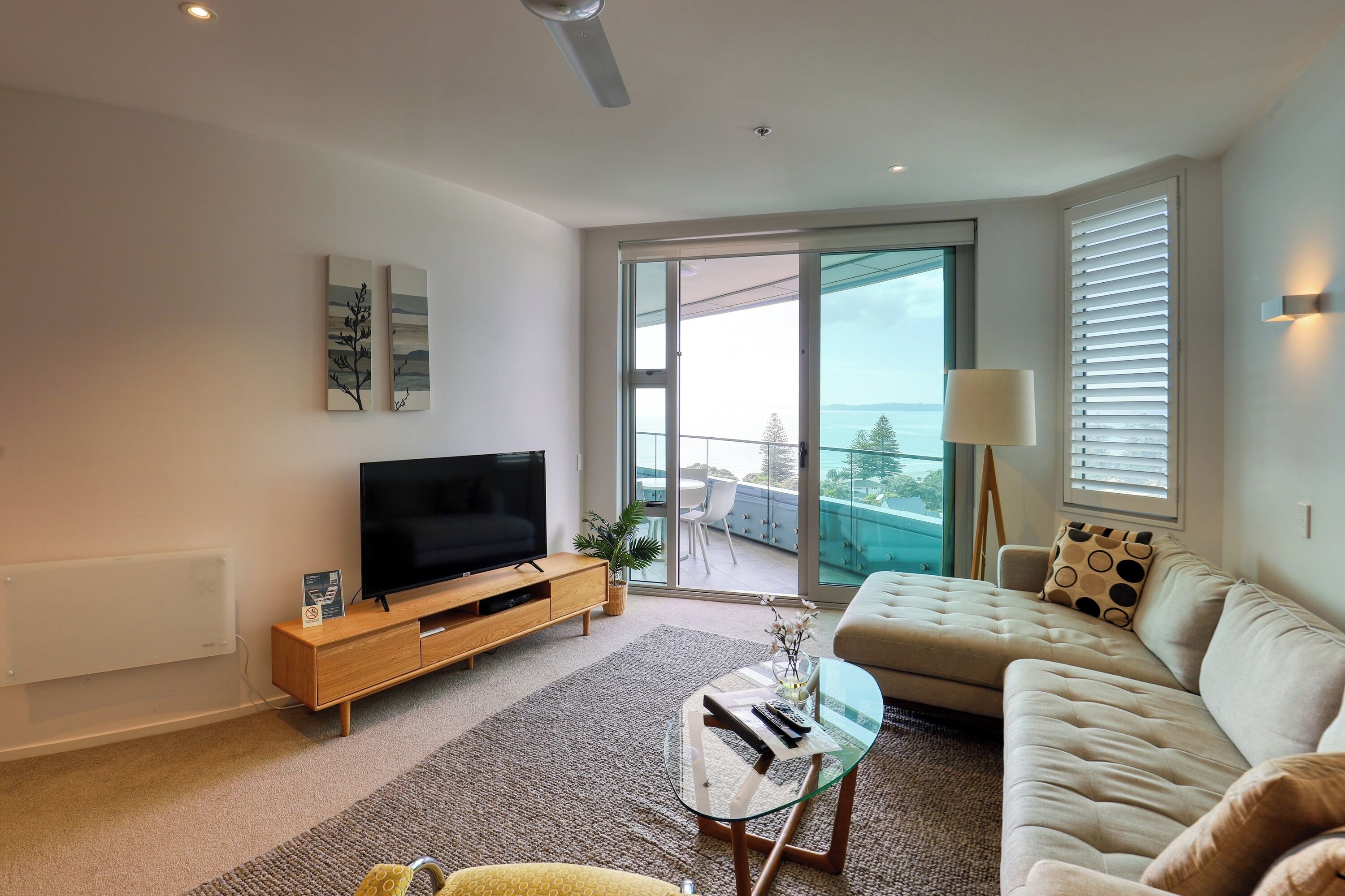 Uma sala de estar moderna com uma TV de tela plana, um sofá, uma mesa de centro de vidro e vista para o exterior por grandes janelas.