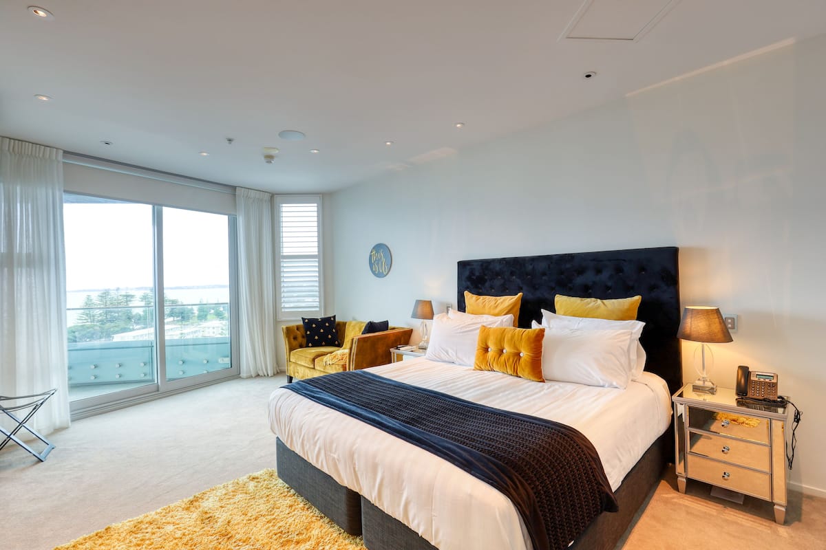 A modern bedroom with a large bed, a nightstand, a chair, and a view of the outdoors through large windows.
