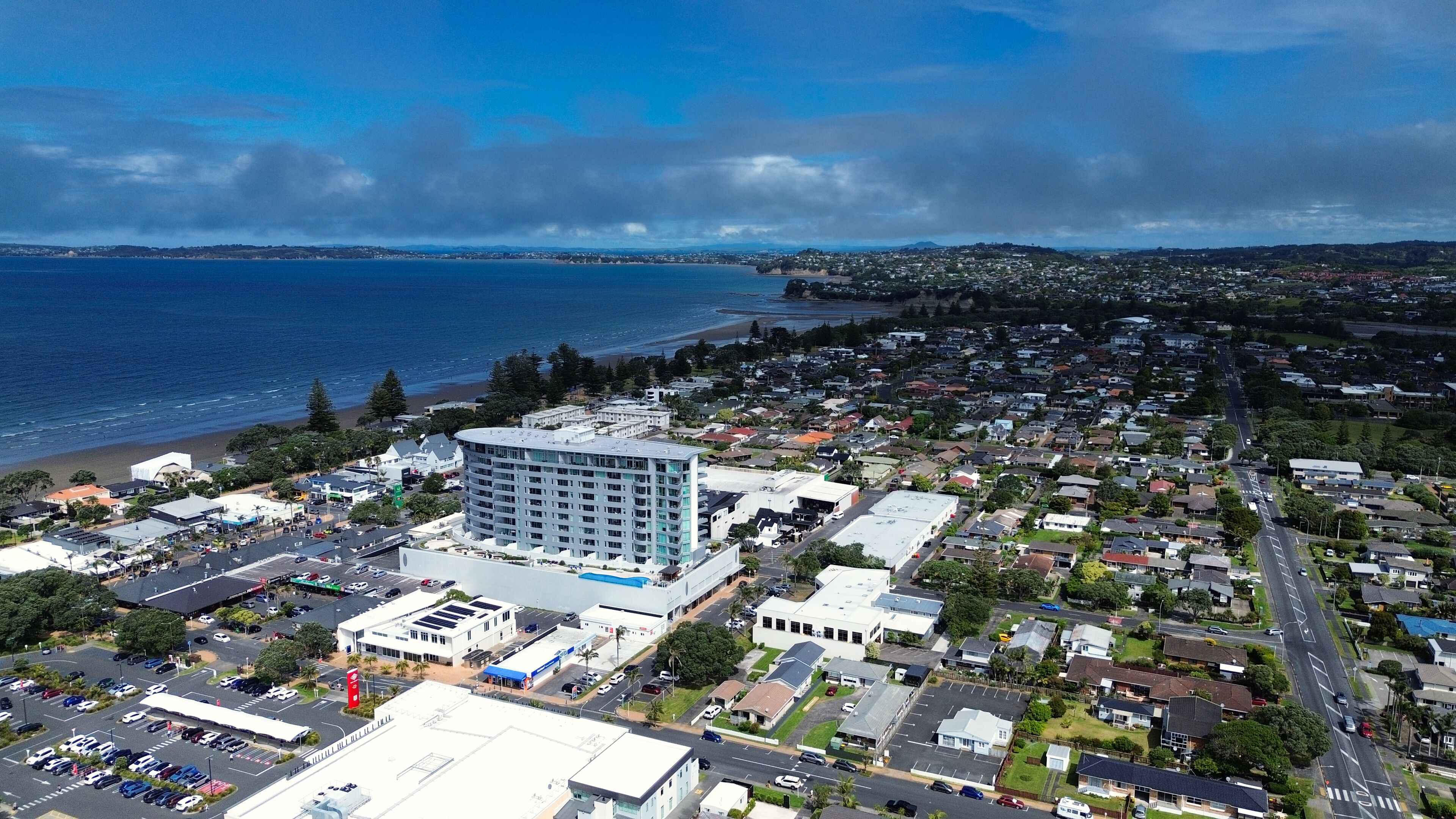 A coastal cityscape with a prominent hotel featuring multiple floors and a rooftop pool, surrounded by residential areas and a beach.