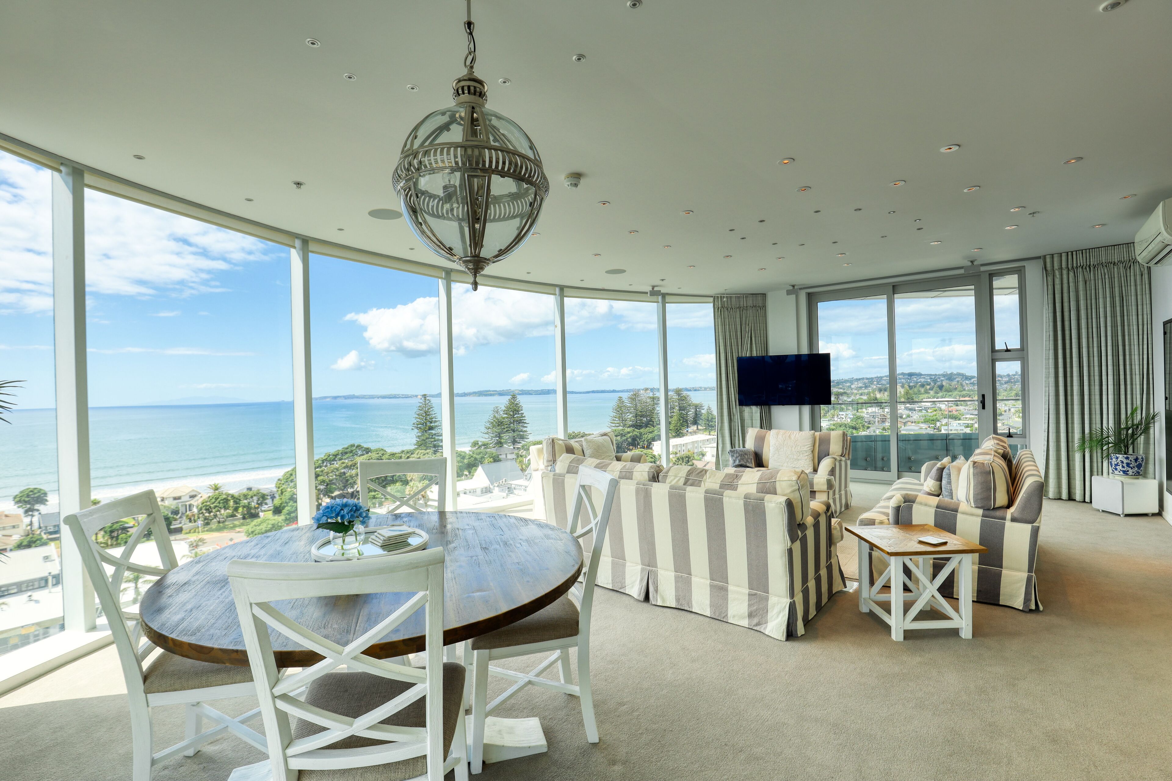 A spacious living room with a large window overlooking the sea, a round wooden table, and striped sofas.