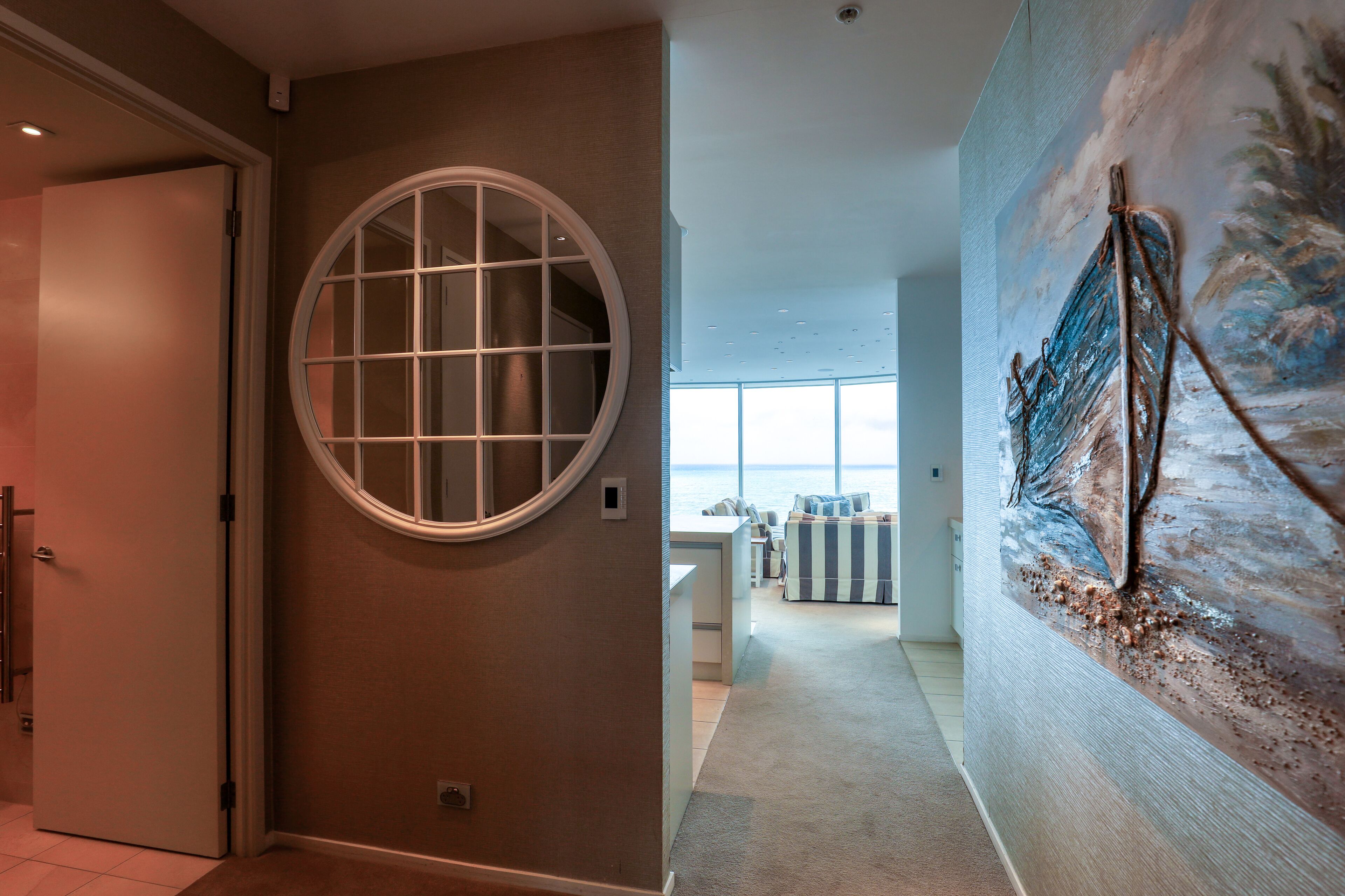 A hallway with a round window, a painting of a shipwreck, and a view of the ocean through large windows.
