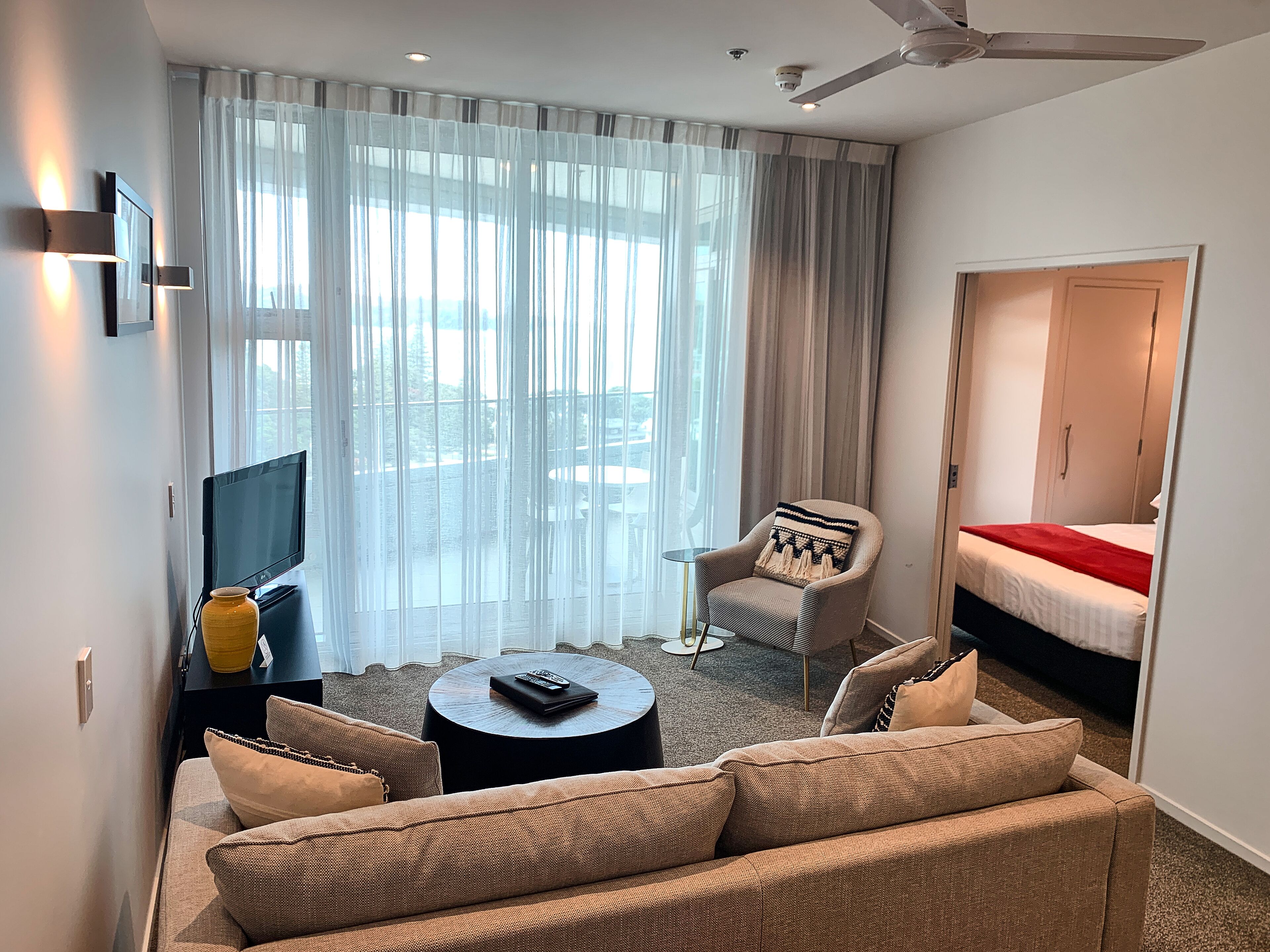 A hotel room with a sofa, armchair, and a bed. There is a TV and a small table with a remote control.