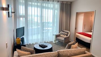 A hotel room with a sofa, armchair, and a bed. There is a TV and a small table with a remote control.