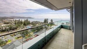 A balcony with a glass railing overlooking a parking area and a distant view of a body of water and hills. - Marsden Suites Nautilus Orewa (Orewa)