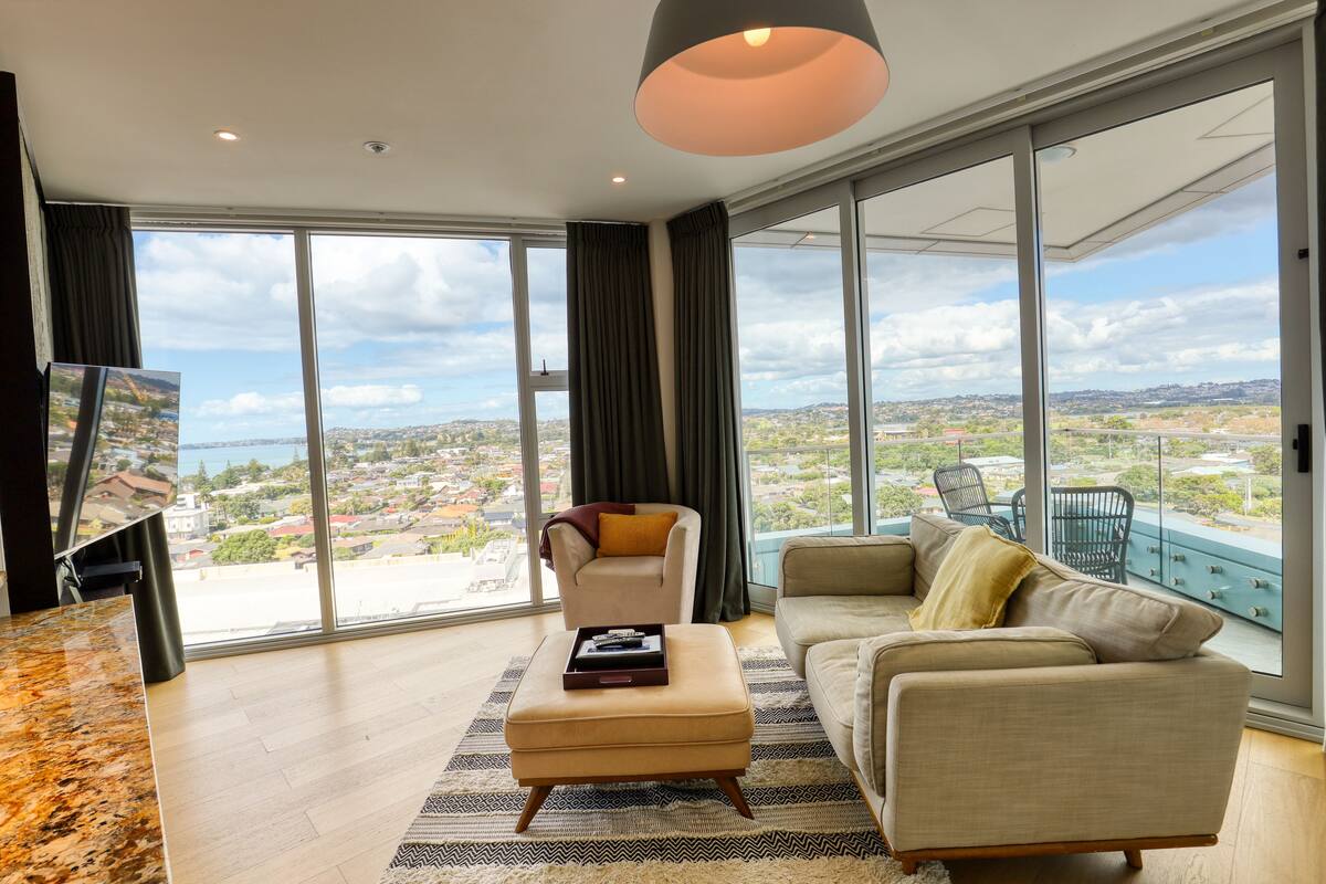 A spacious living room with a large window offering a city view, a comfortable sofa, a chair, and a small ottoman with a tray.