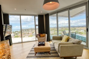 A spacious living room with a large window offering a city view, a comfortable sofa, a chair, and a small ottoman with a tray. - Marsden Suites Nautilus Orewa (Orewa)