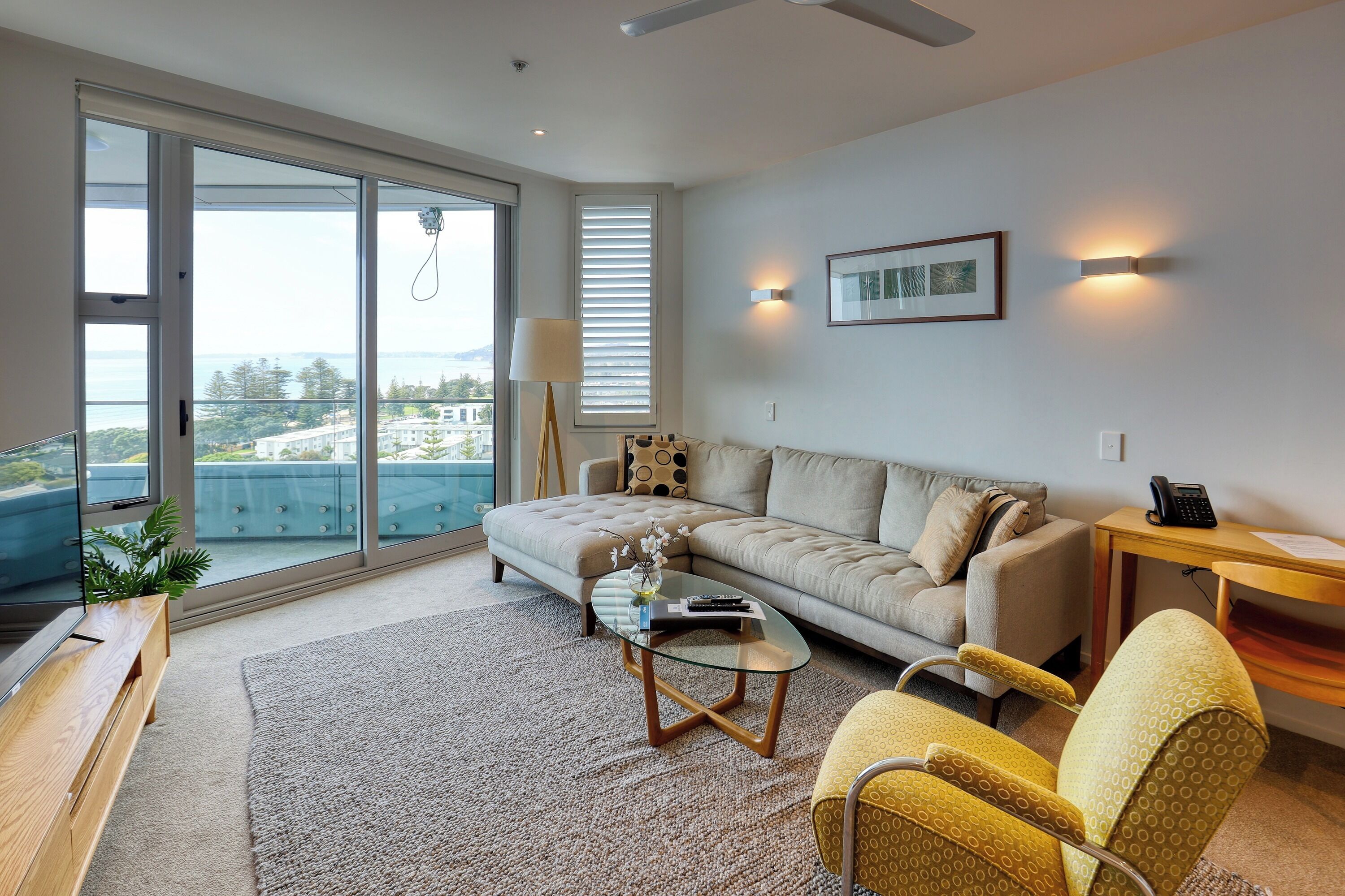 A modern living room with a sofa, yellow armchairs, a glass coffee table, and a view of the city through large windows.