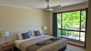 Un dormitorio con cama, mesitas de noche, ventilador de techo y ventana con vista a la vegetación.