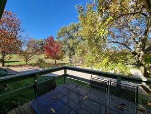 A covered outdoor seating area with a metal table and chairs, overlooking a park with trees and a clear sky.