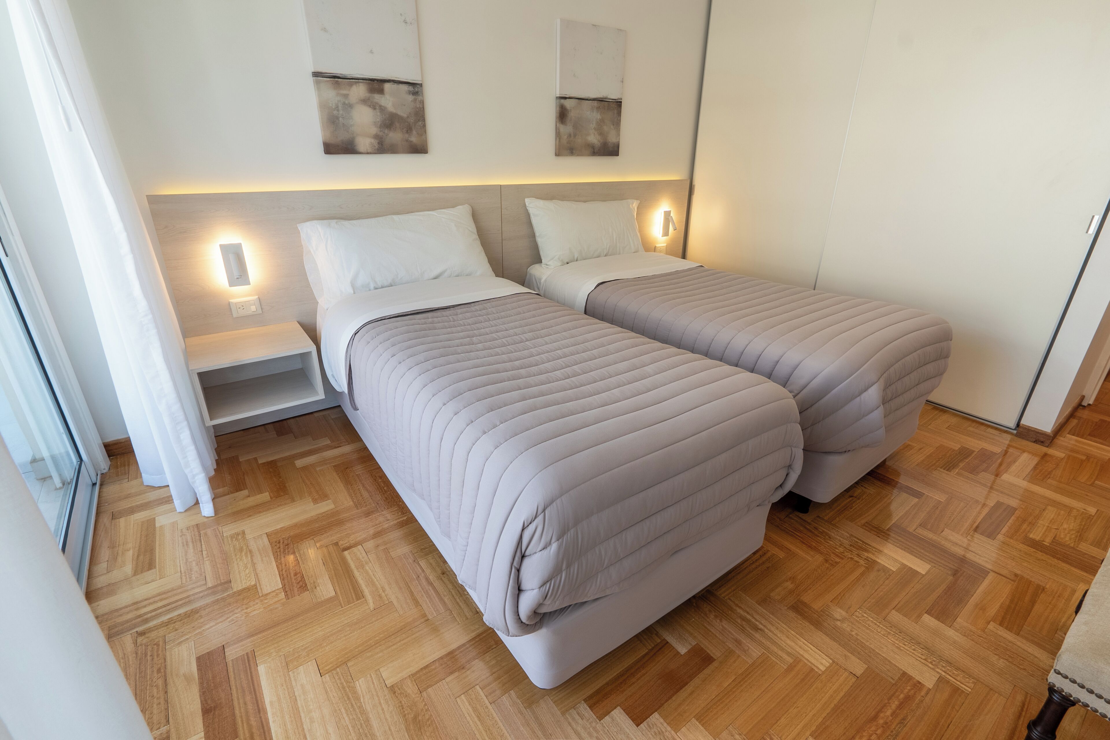 Una habitación de hotel con dos camas, suelo de parqué y lámparas fijadas en la pared.
