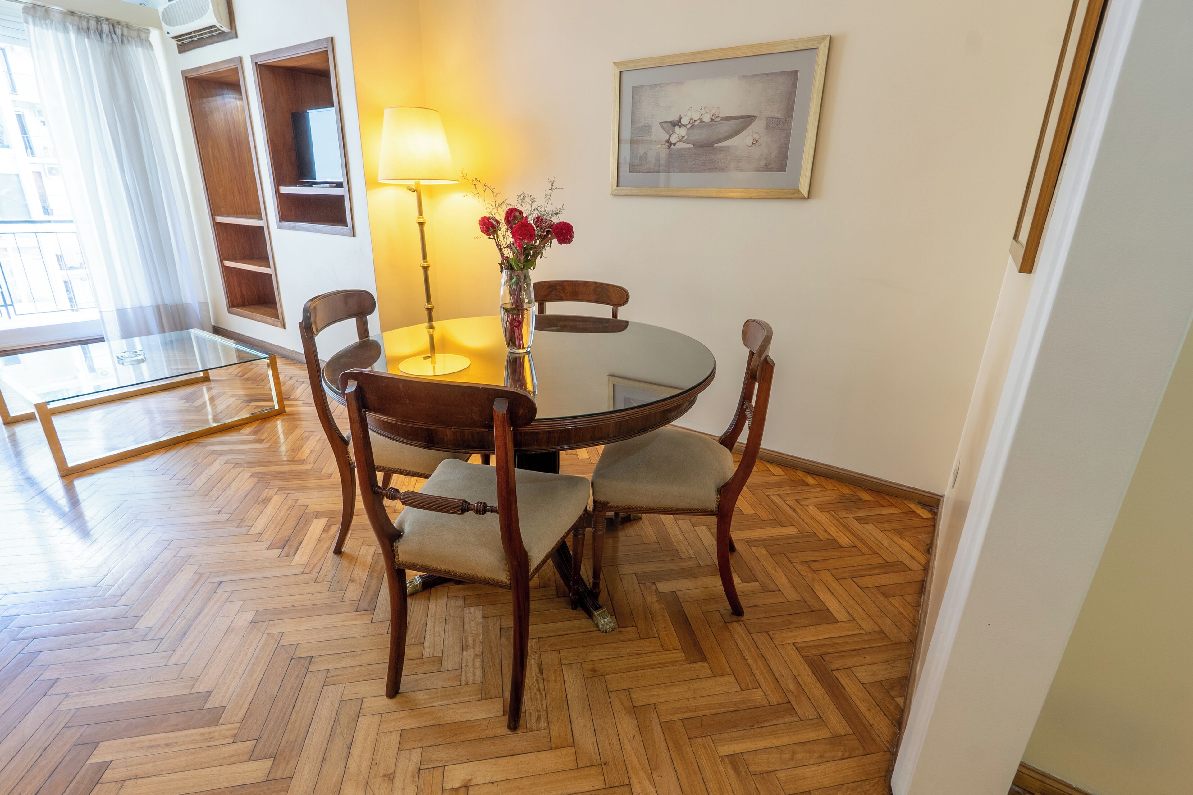 A dining area with a round table, four chairs, a vase with flowers, a lamp, and a framed picture on the wall.
