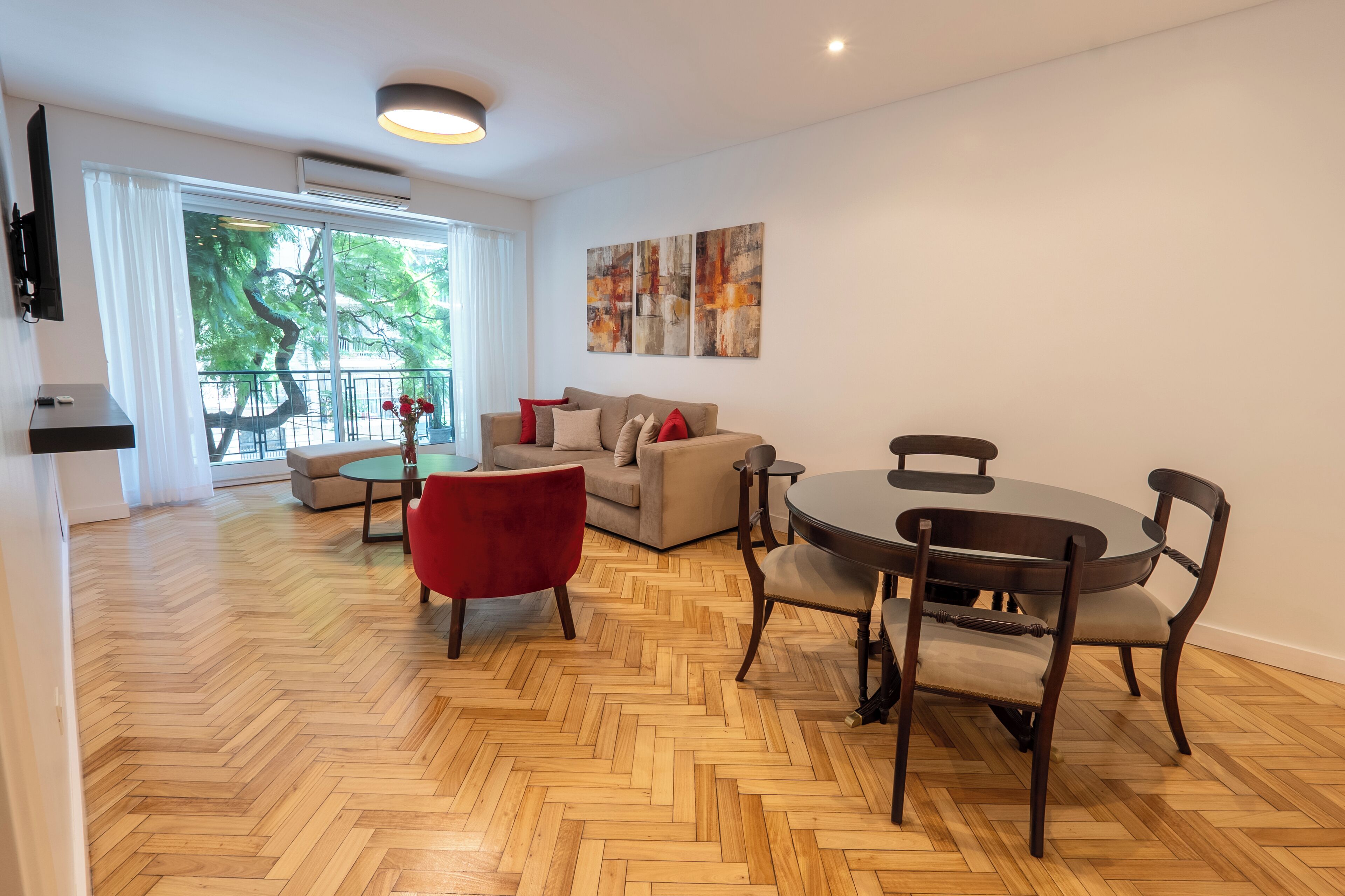 A spacious living area with a herringbone wooden floor, a beige sofa, a red chair, a round dining table with four chairs, and a large window with a view of trees.