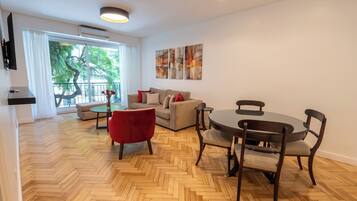 A spacious living area with a herringbone wooden floor, a beige sofa, a red chair, a round dining table with four chairs, and a large window with a view of trees.