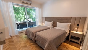 Habitación doble con dos camas, piso de madera en espiga, un ventanal grande con cortinas y un balcón con árboles.
