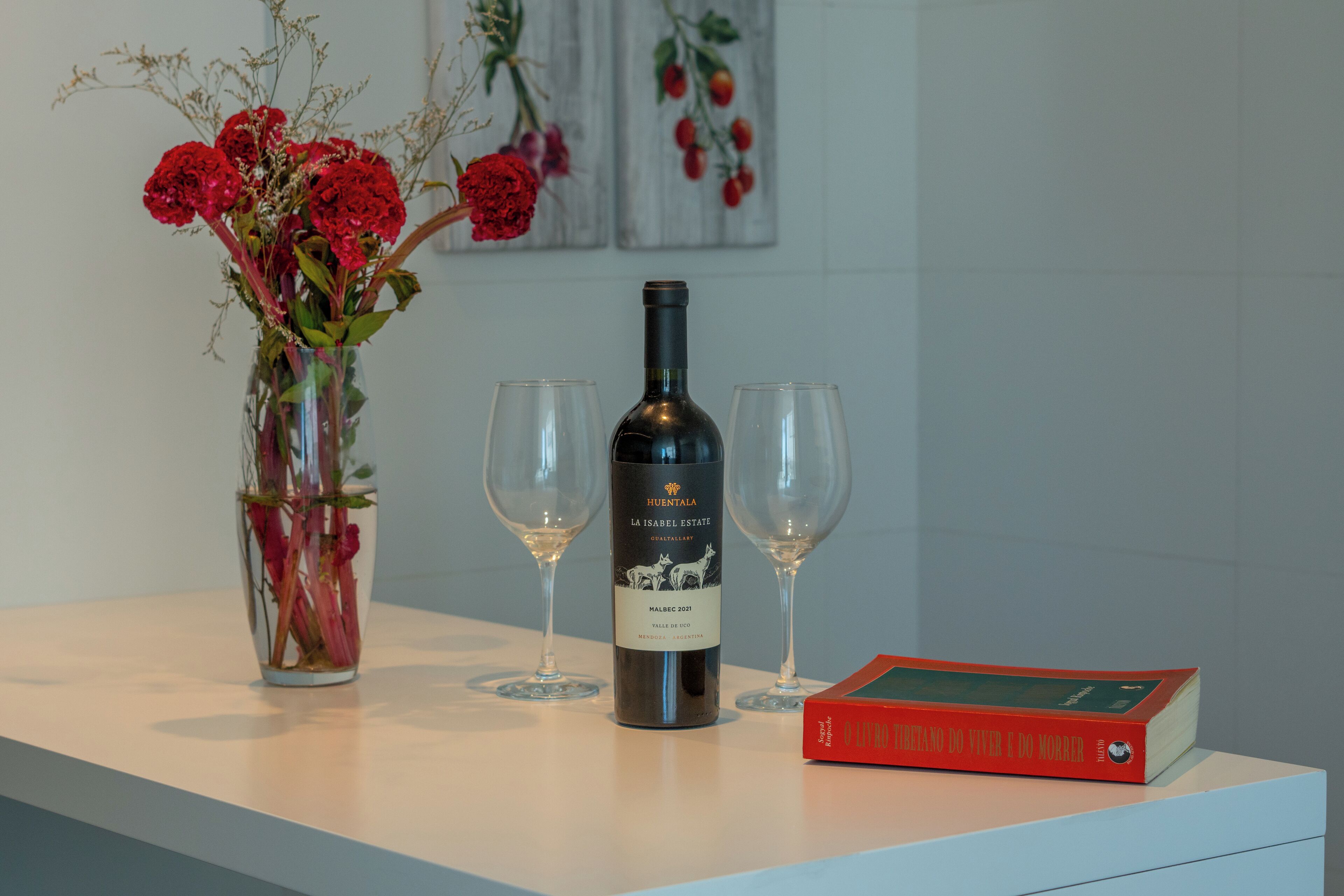 A wine bottle, two wine glasses, a vase with flowers, and a book on a table.