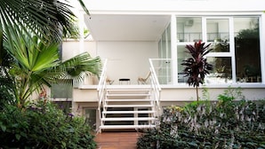 Un edificio moderno con fachada blanca, una escalera de madera que conduce a un balcón y abundante vegetación alrededor de la entrada.