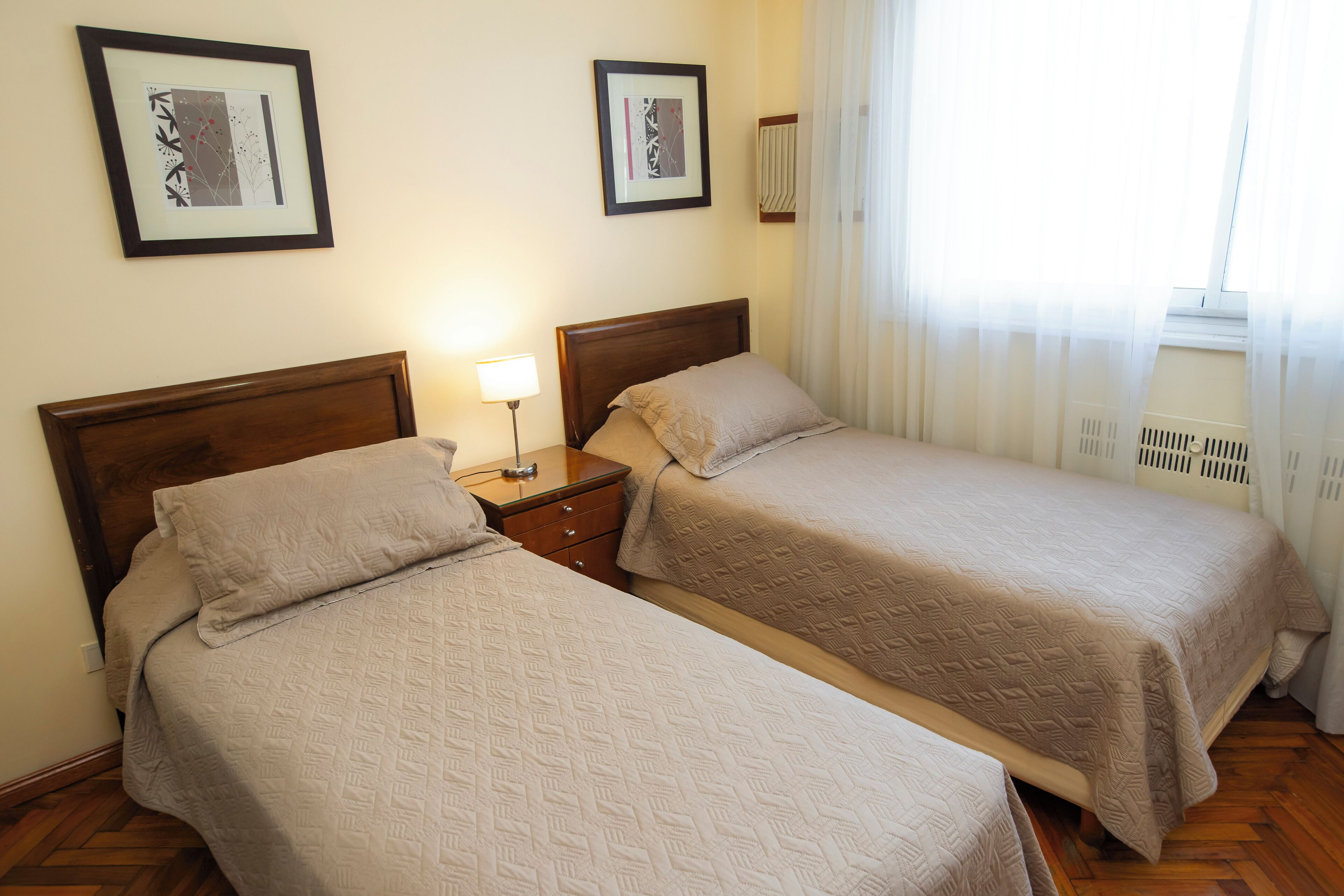 A hotel room with two beds, a nightstand with a lamp, and framed artwork on the walls.