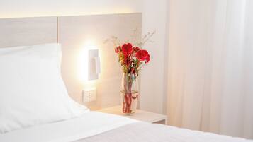 A neatly made bed with white linens, a bedside table with a vase of red flowers, and a wall-mounted light.